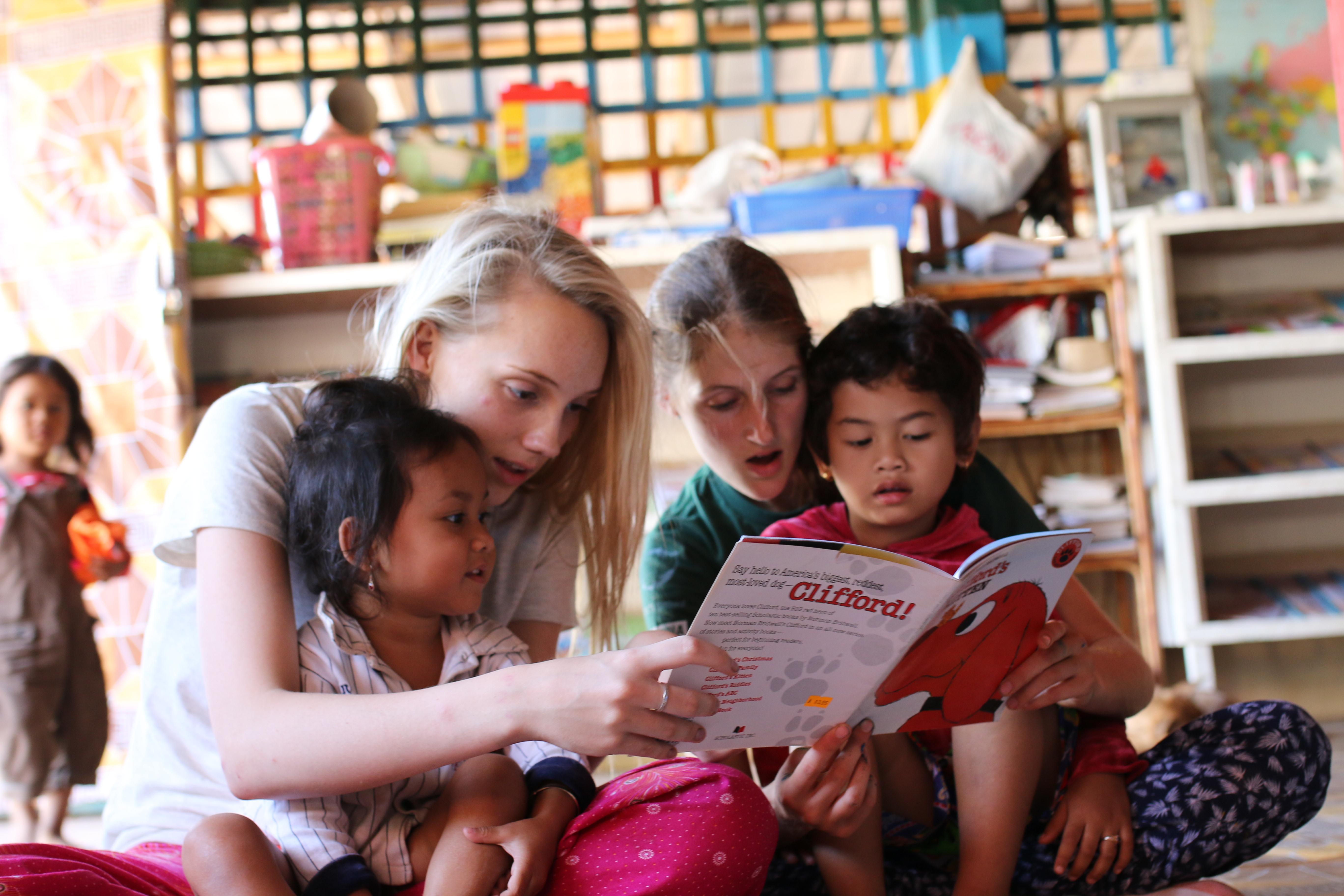 Two female Projects Abroad Childcare volunteers read a book to children at a day care centre in Cambodia. Children at a day care centre listen to a book being read by two female volunteers working with children in Cambodia.